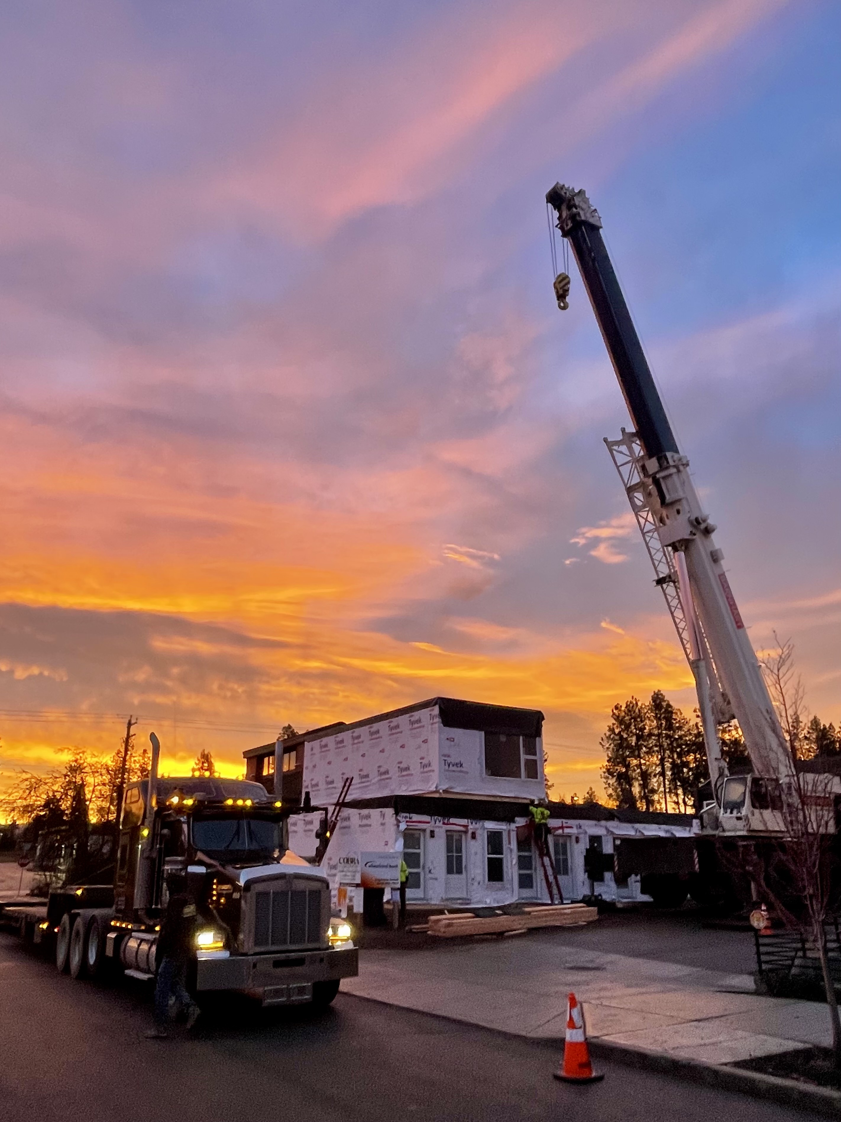 Crane and flatbed truck moving a modular building unit at sunset on a DanReDev site